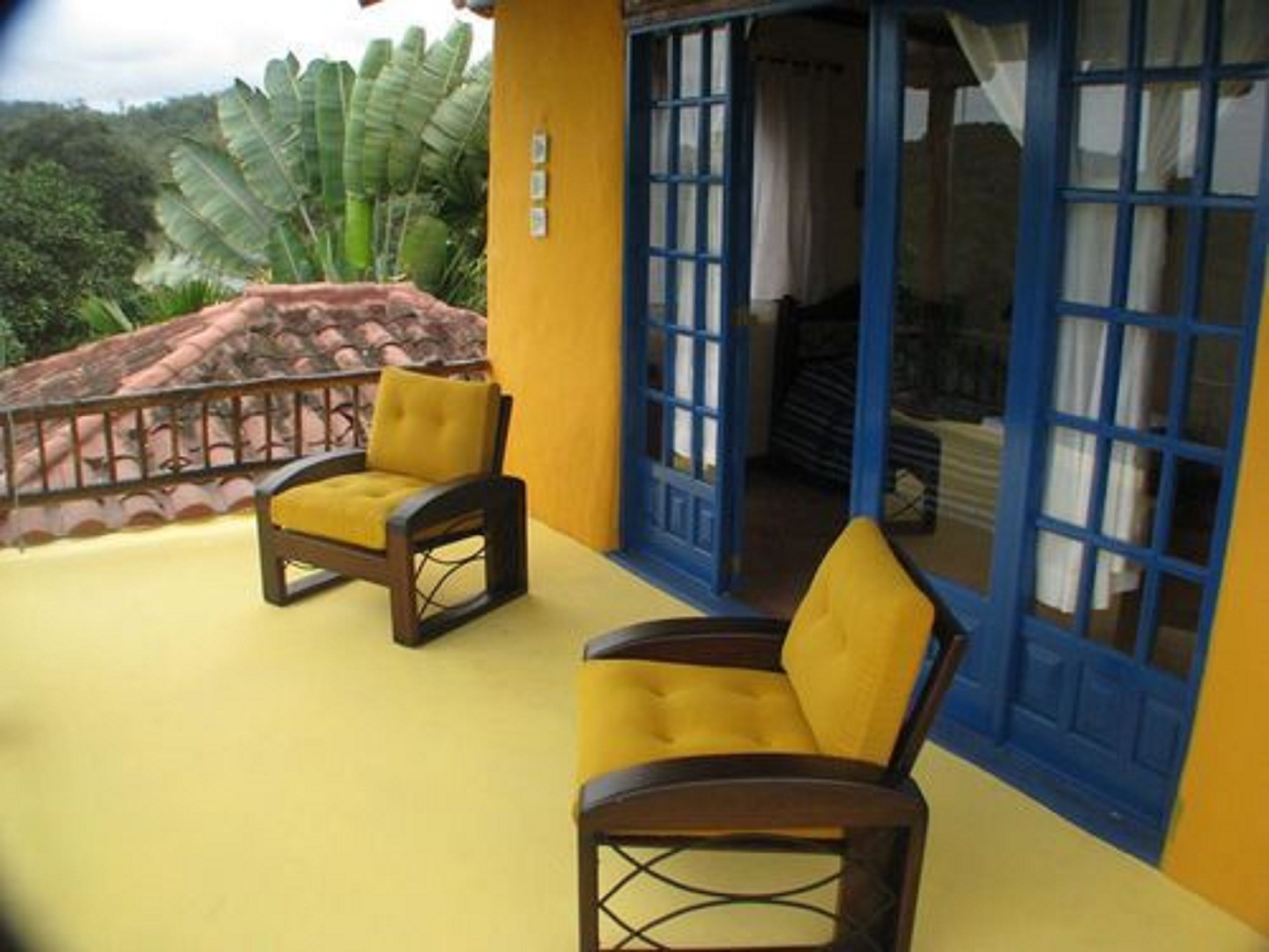 Un edificio amarillo con puertas y ventanas azules, dos sillas amarillas afuera y vistas a follaje verde.