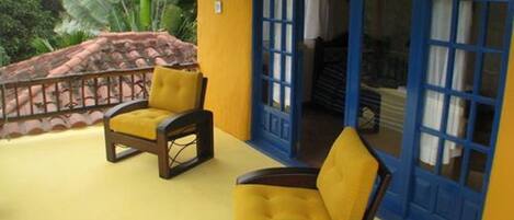Un edificio amarillo con puertas y ventanas azules, dos sillas amarillas afuera y vistas a follaje verde.