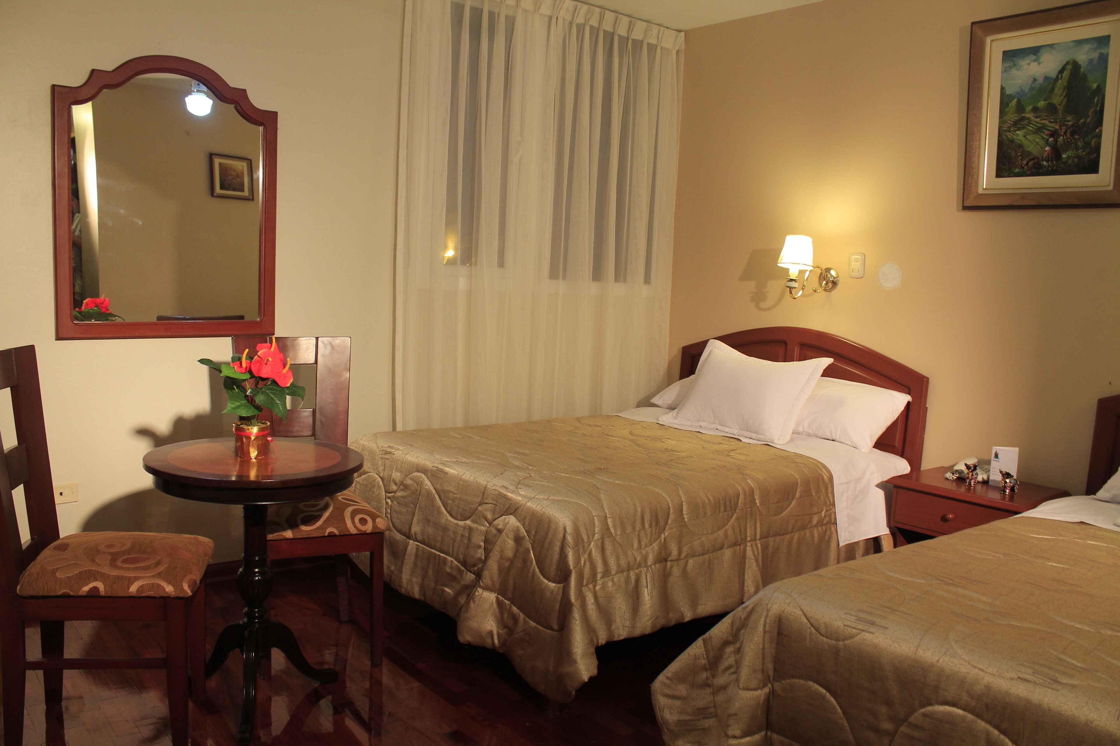 Habitación de hotel con dos camas, una silla de madera, una mesita redonda con un jarrón de flores, un espejo y un cuadro en la pared.