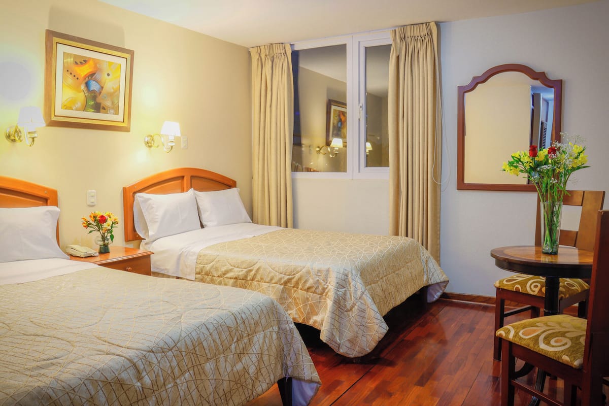 Une chambre d’hôtel avec deux lits, une fenêtre avec des rideaux, un miroir, une petite table sur laquelle est posé un vase de fleurs, et un tableau accroché au mur.