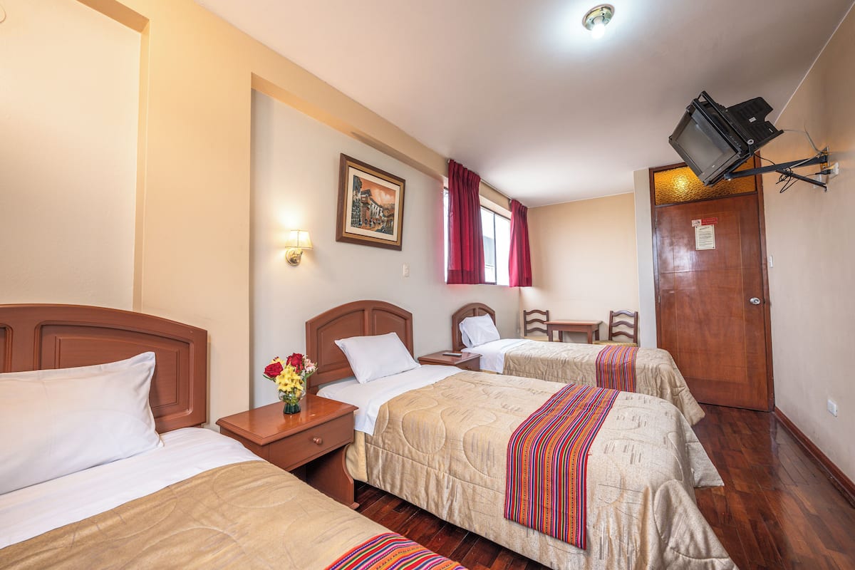 Une chambre d’hôtel avec trois lits, une tête de lit en bois, une table de chevet ornée d’un vase de fleurs, un tableau encadré accroché au mur et un téléviseur fixé au mur.