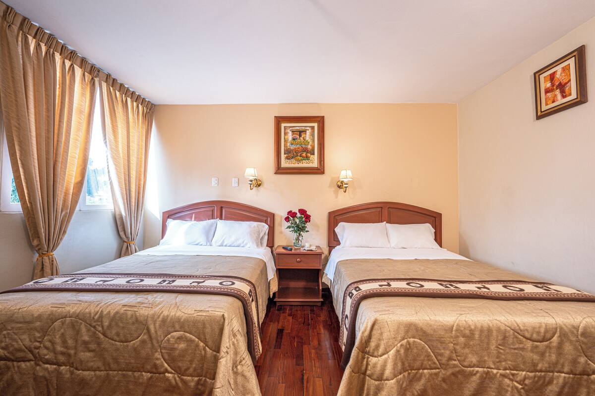 Une chambre d’hôtel avec deux lits, des têtes de lit en bois, des murs de couleur beige, un tableau encadré au-dessus des lits et une table de chevet avec un vase de fleurs placé entre les lits.