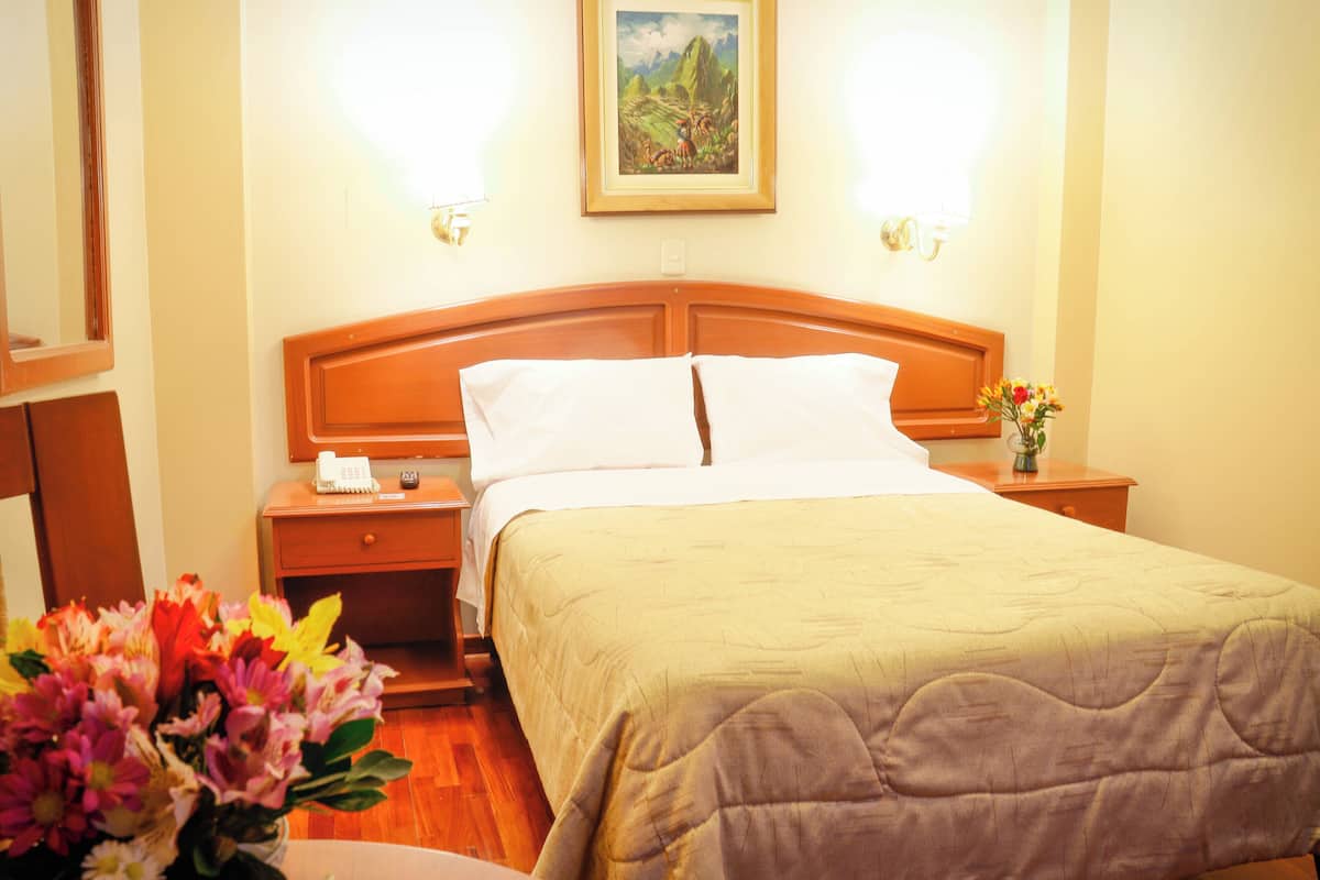 Une chambre d’hôtel avec une tête de lit en bois, un lit recouvert d’une courtepointe beige, une table de chevet avec un téléphone, un vase de fleurs et un tableau au mur.