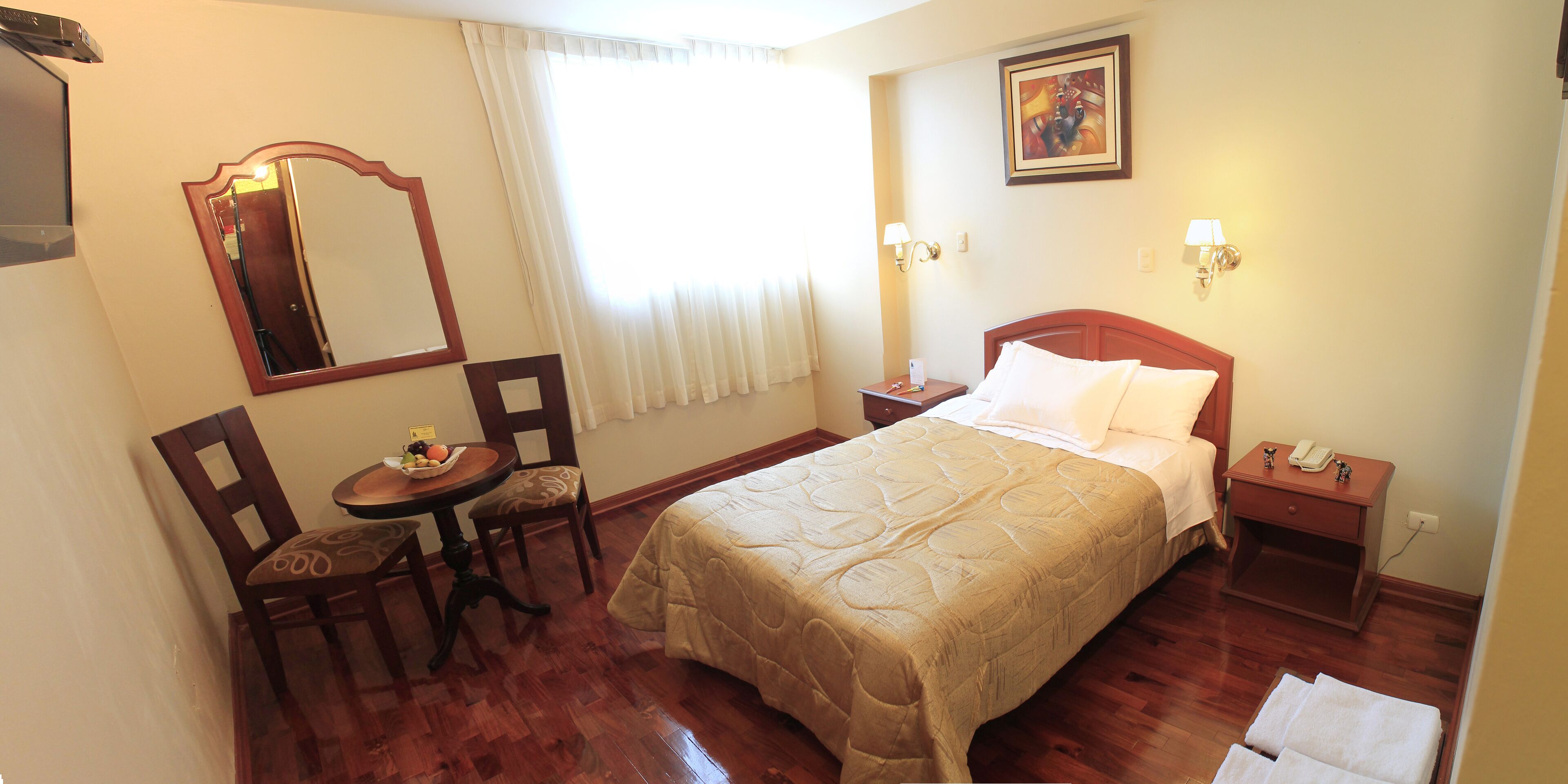 Habitación de hotel con cama, escritorio y silla, una mesita con fruta, espejo y un cuadro en la pared.