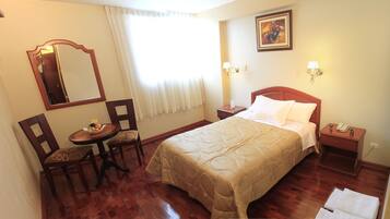 Habitación de hotel con cama, escritorio y silla, una mesita con fruta, espejo y un cuadro en la pared.
