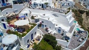Un edificio blanco con puertas y ventanas azules, rodeado de vegetación y otros edificios.