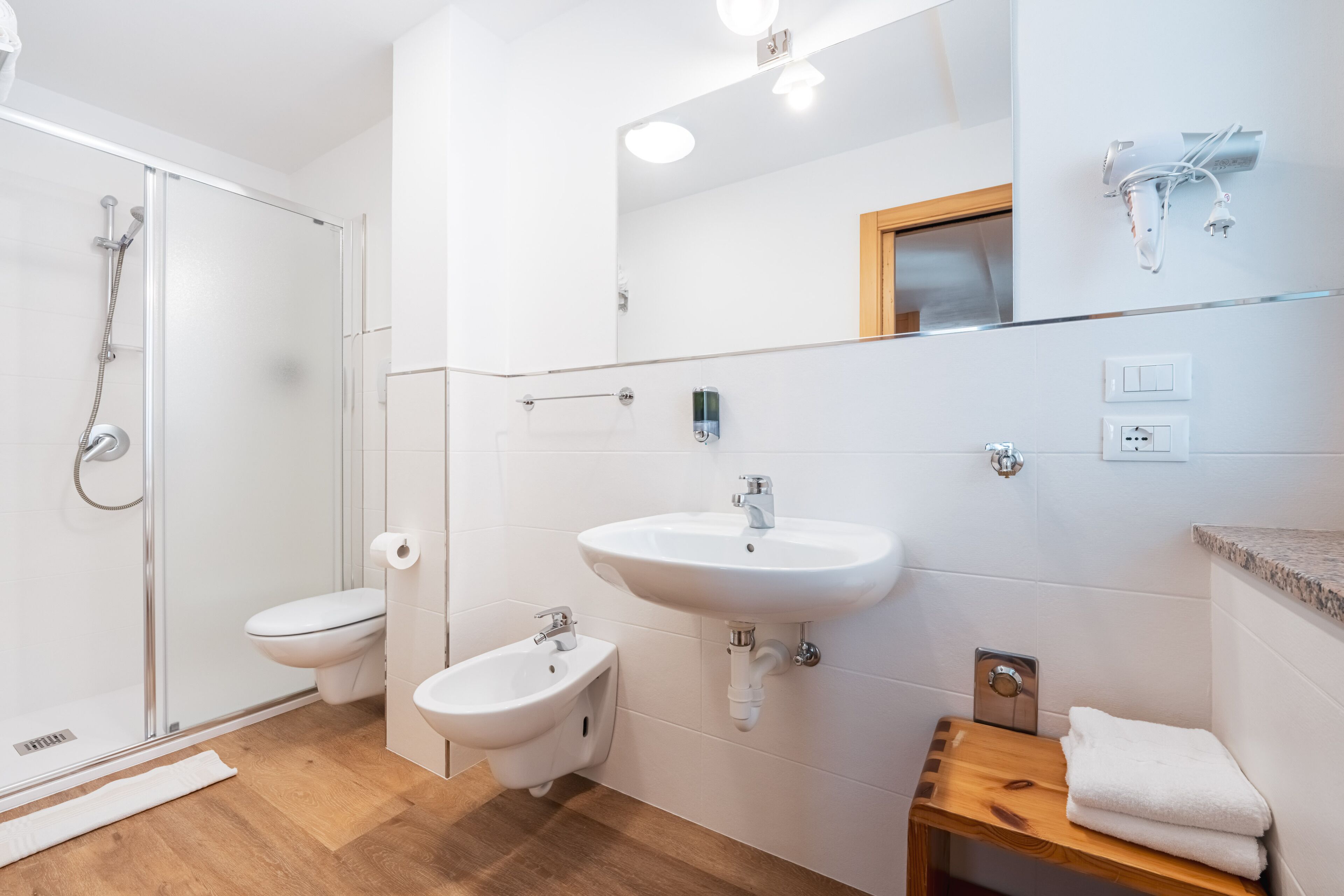 A modern bathroom with a glass shower enclosure, a toilet, a bidet, a sink, and a wooden shelf with towels.