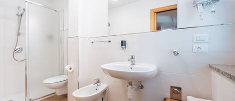 A modern bathroom with a glass shower enclosure, a toilet, a bidet, a sink, and a wooden shelf with towels.