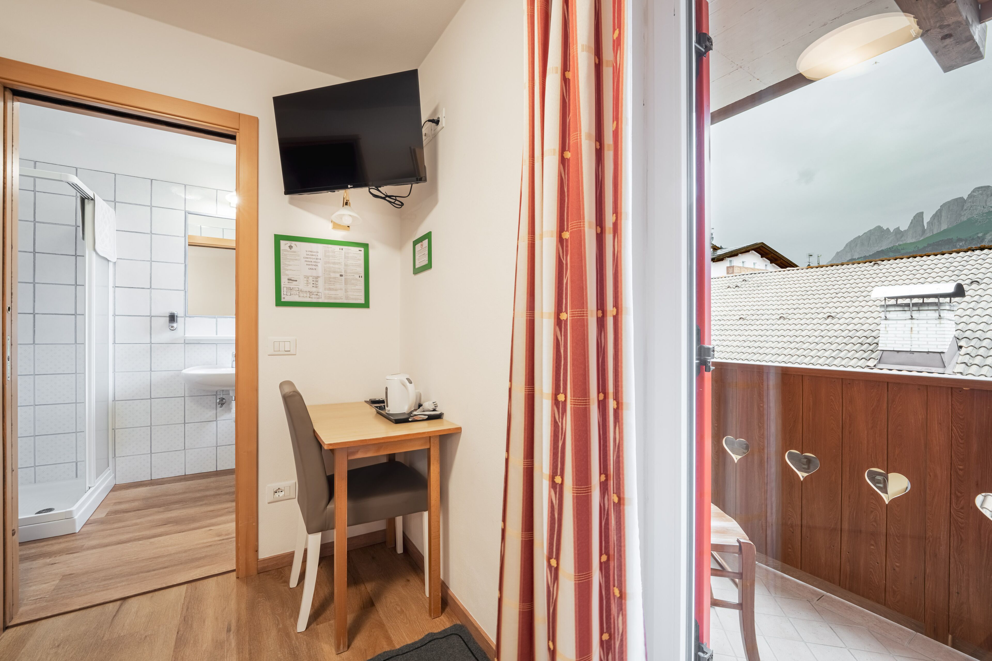 Habitación compacta con ducha, una pequeña mesa con silla, televisor montado y balcón con baranda de madera y recortes en forma de corazón.