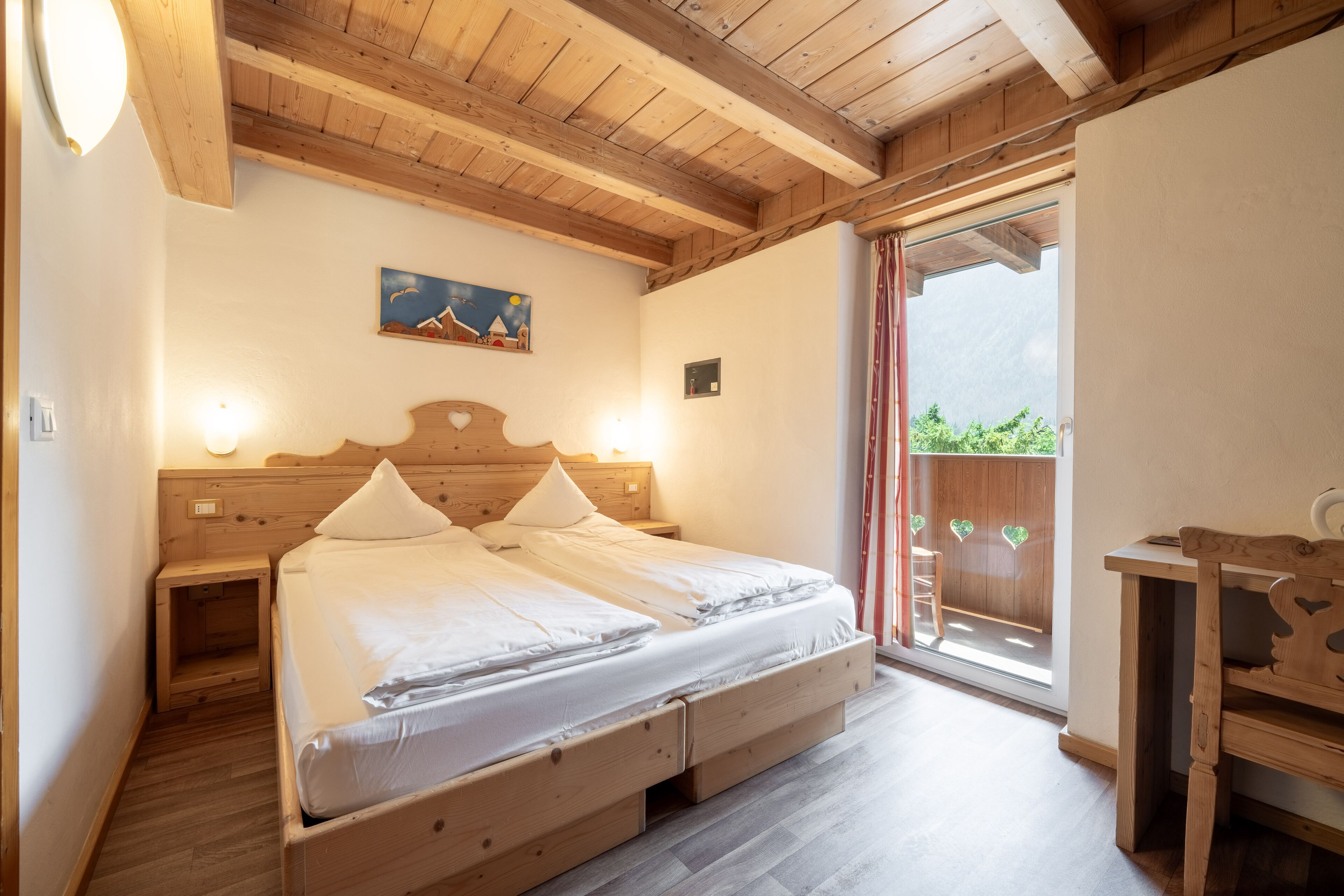 A wooden-framed bedroom with a bed, bedside tables, a painting, and a view of greenery through a sliding door.