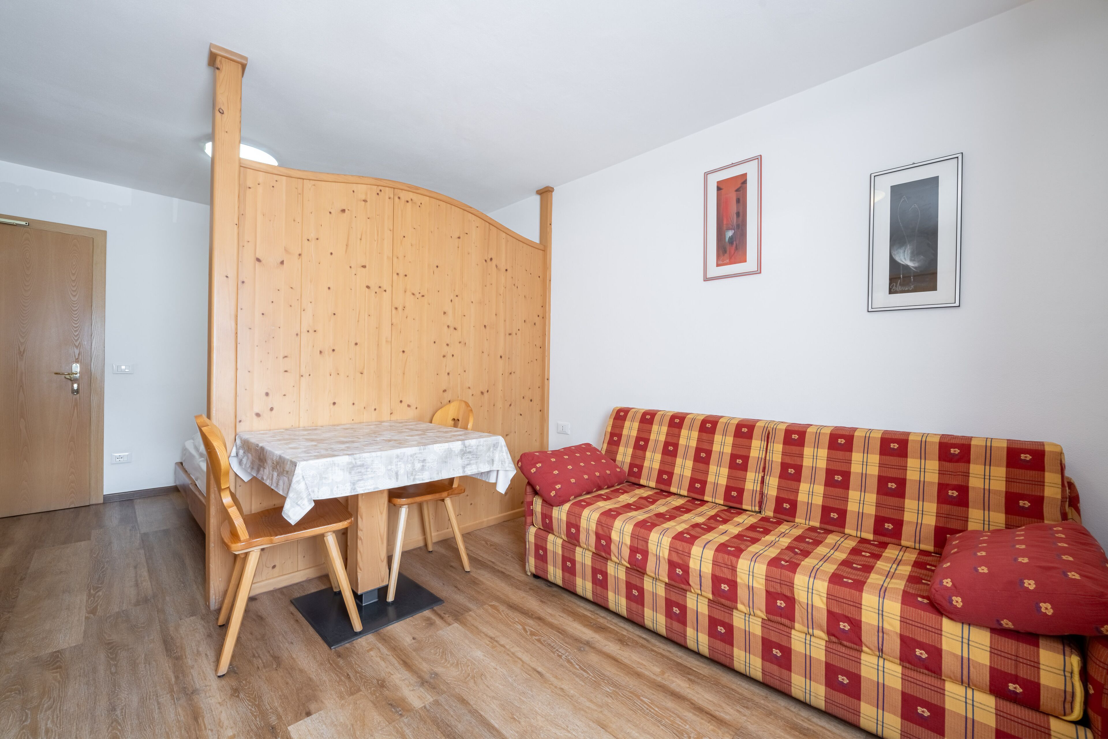A room with a wooden dining table and chairs, a plaid sofa, and two framed pictures on the wall.