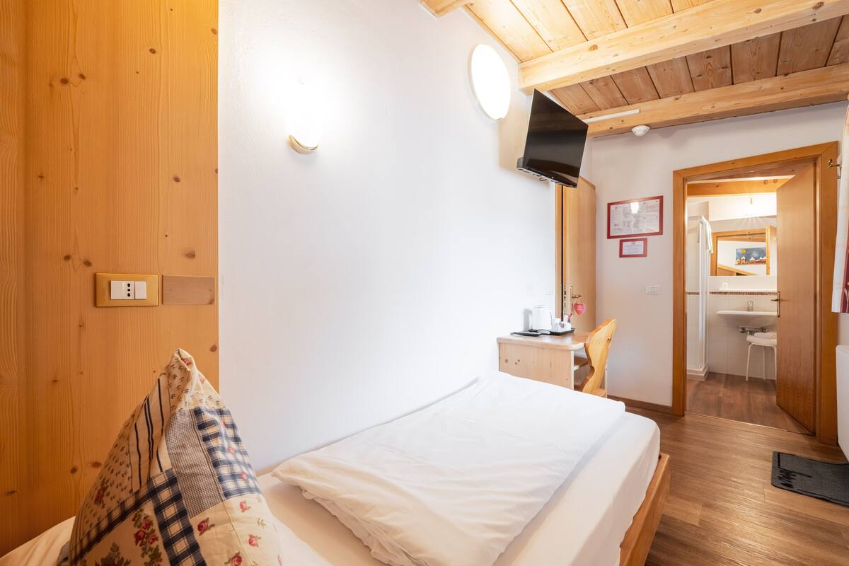 Makuuhuone, jossa on puinen katto, valkoisilla lakanoiden peitetty sänky, yöpöytä lampun kanssa ja näkymä kylpyhuoneeseen.