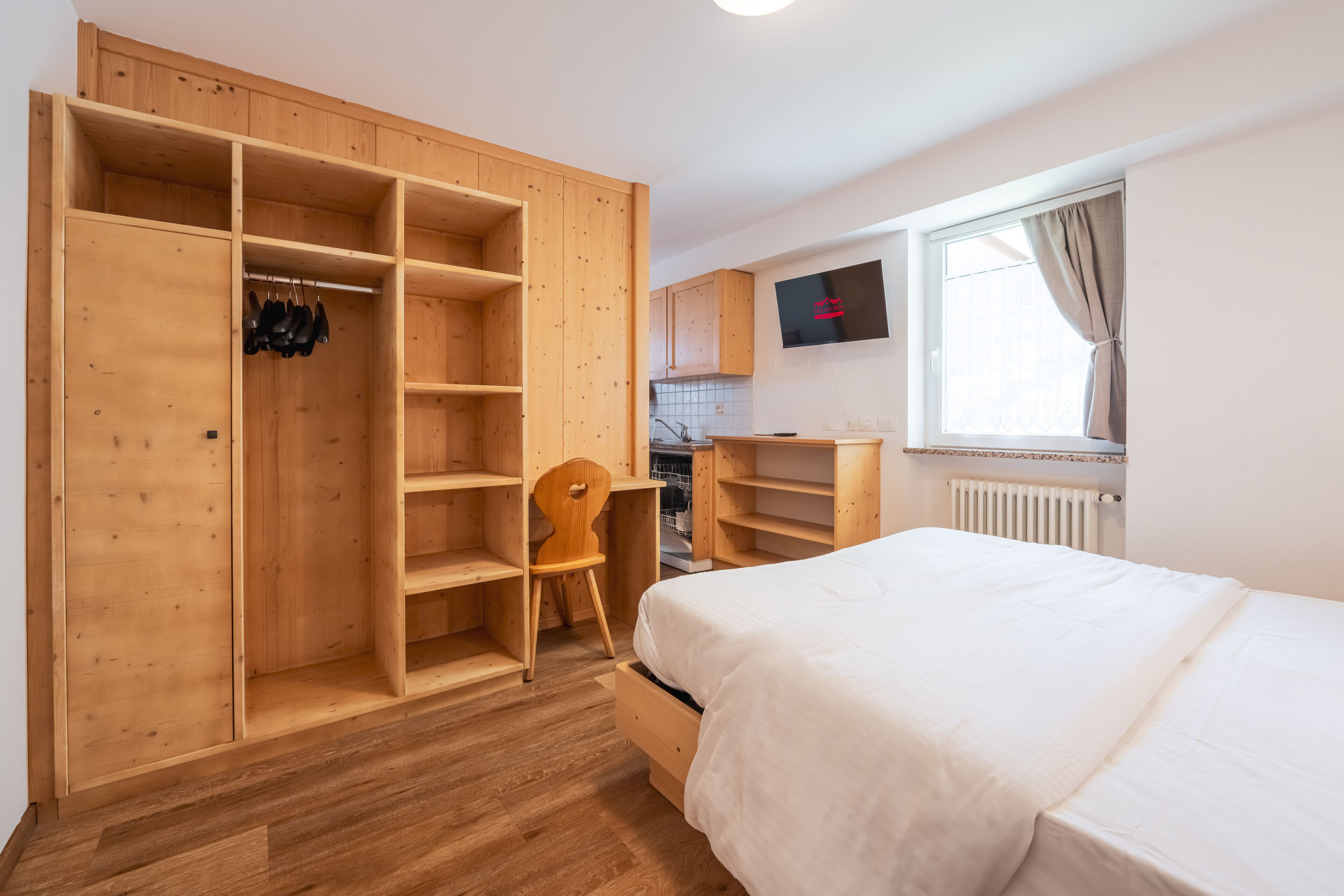 A bedroom with a wooden wardrobe, a bed, a wooden chair, and a kitchen area with a TV.