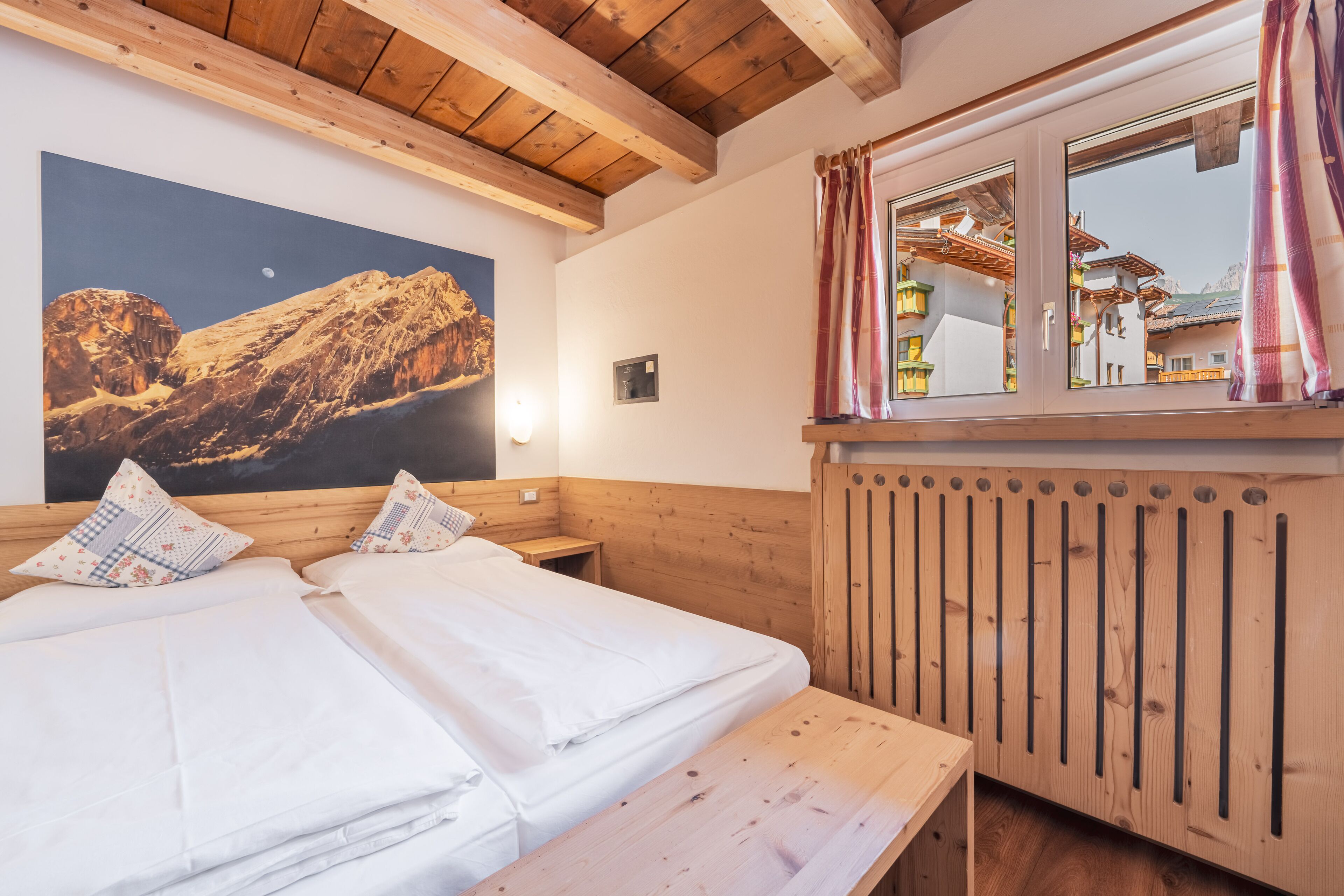 A room with two beds, wooden beams, a large mountain painting, a window with curtains, and a wooden bench.