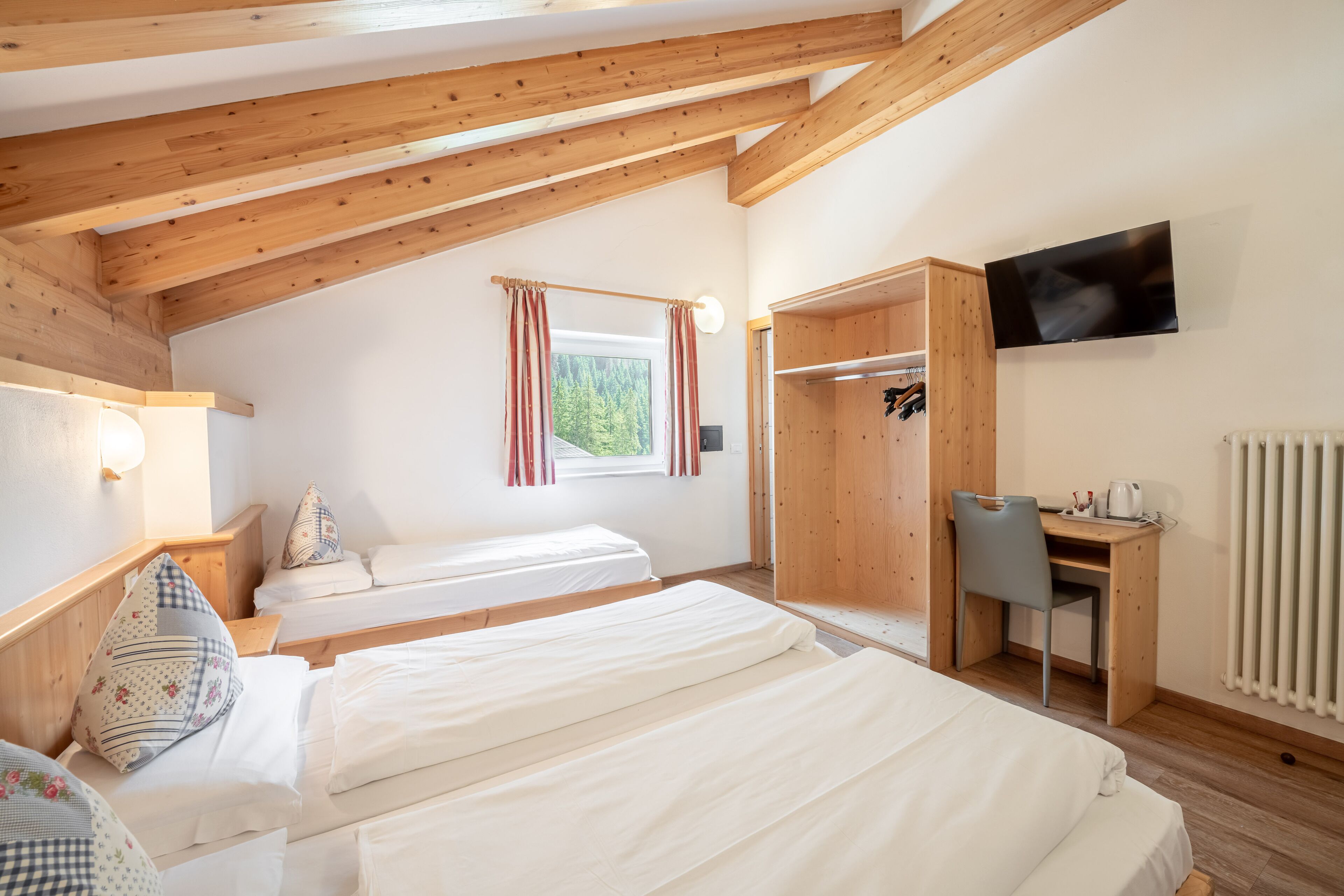 Quarto com duas camas, uma escrivaninha de madeira, uma cadeira, uma televisão e uma janela com cortinas.