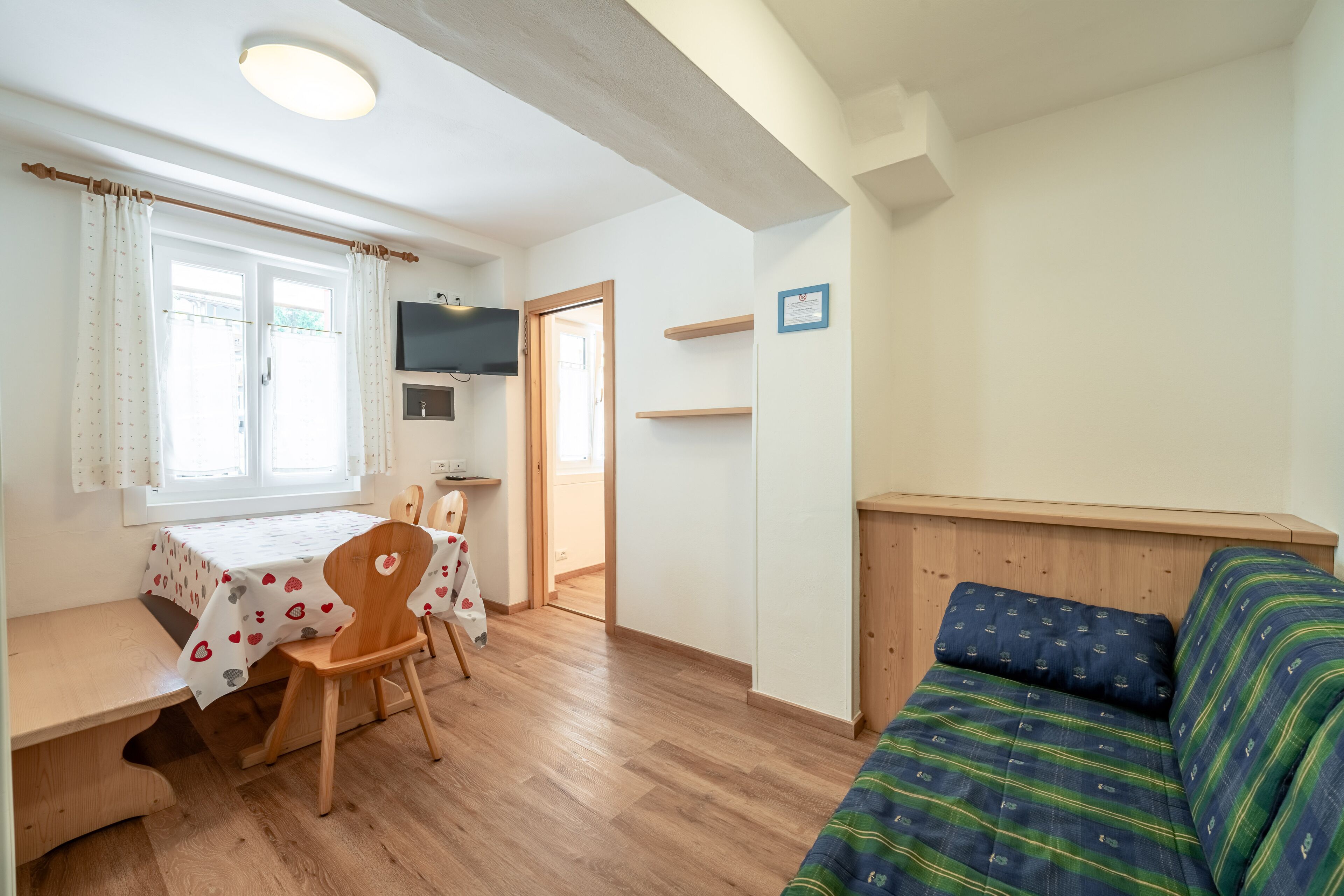 Een compacte kamer met een houten vloer, een eettafel met een tafelkleed met hartjesprint, een houten stoel, een bank met een gestreept kussen, een televisie aan de muur en een deur die leidt naar een andere kamer.