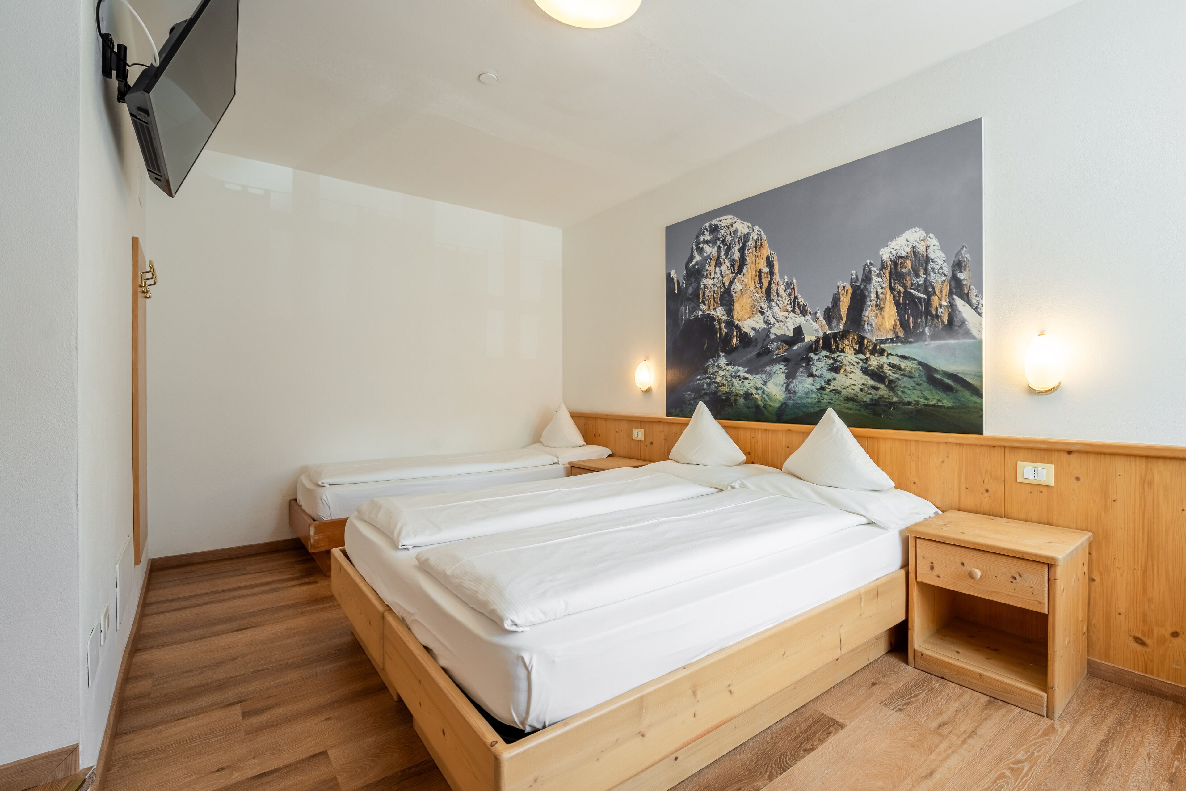 Un dormitorio con dos camas de madera, una mesita de noche, un televisor montado y un cuadro de un paisaje montañoso.