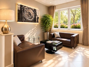 A modern living room with a sofa, a coffee table, a lamp, a large wall clock, a potted plant, and a window with a view of trees and flowers. - Hotel Charles Central - Czech Leading Hotels (Prague)