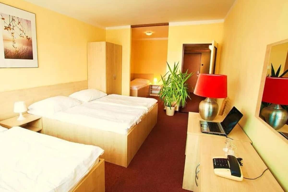 A hotel room with two beds, a desk with a laptop, a red lamp, and a plant.
