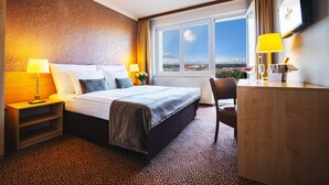 A hotel room with a bed, a desk, a chair, a TV, and a window with a city view. - Hotel Charles Central - Czech Leading Hotels (Prague)