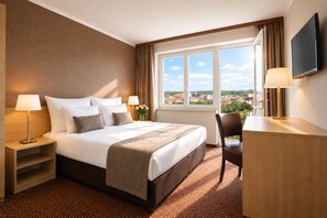 A hotel room with a large bed, two bedside tables with lamps, a desk with a chair, a TV, and a window with a view of buildings. - Hotel Charles Central - Czech Leading Hotels (Prague)