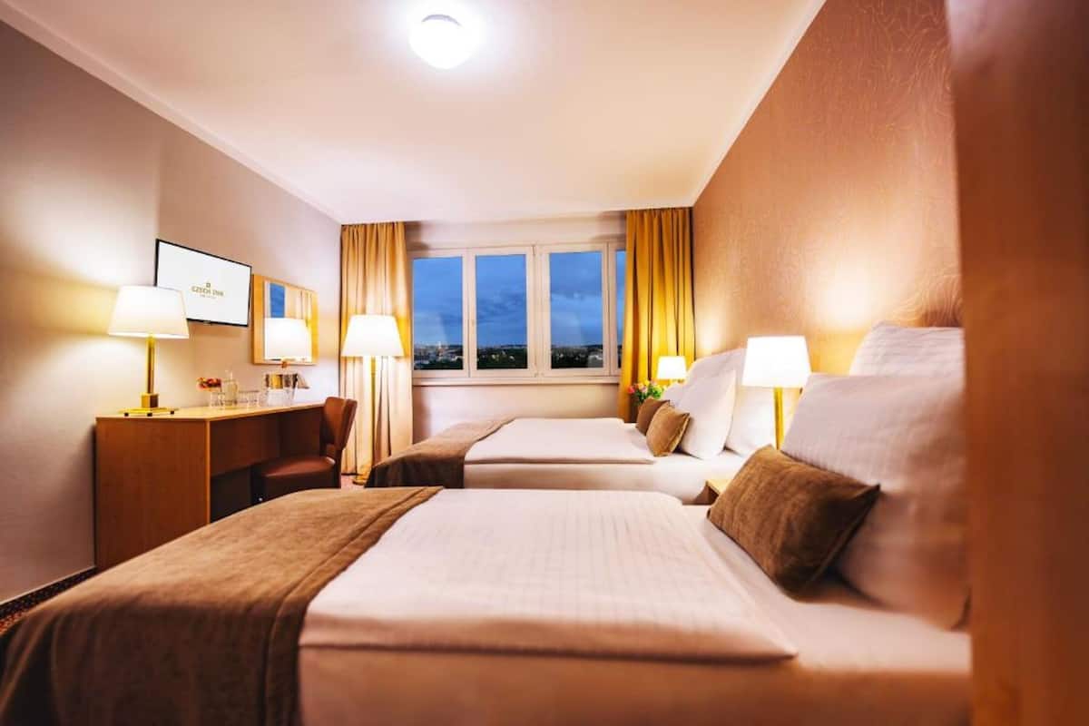 A hotel room with two beds, a desk, and a chair. There is a window with curtains and a view of the cityscape.