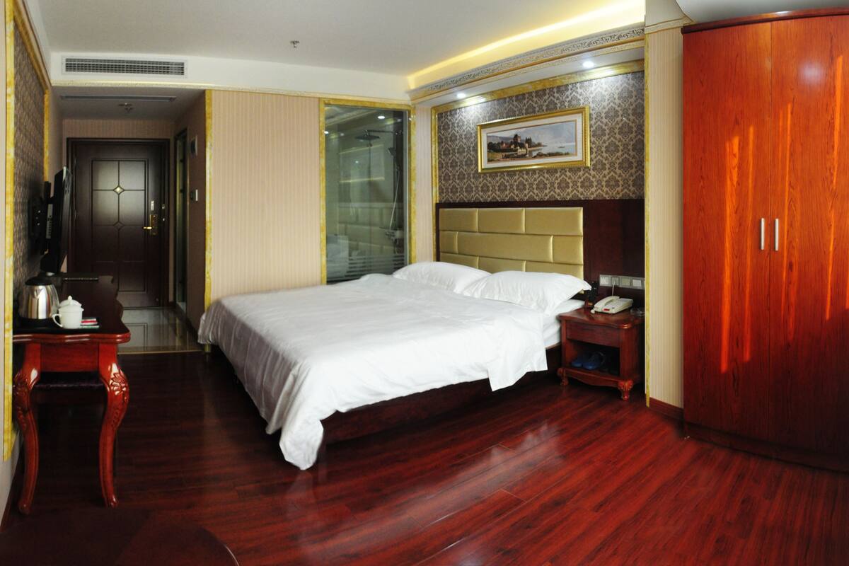 A hotel room with a large bed, a wooden wardrobe, a bedside table with a telephone, and a picture on the wall.