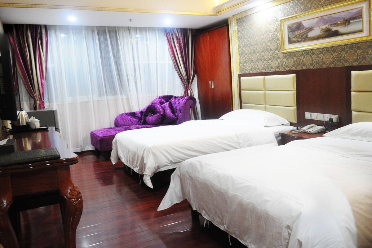 A hotel room with two beds, a desk, a purple armchair, and a painting on the wall.