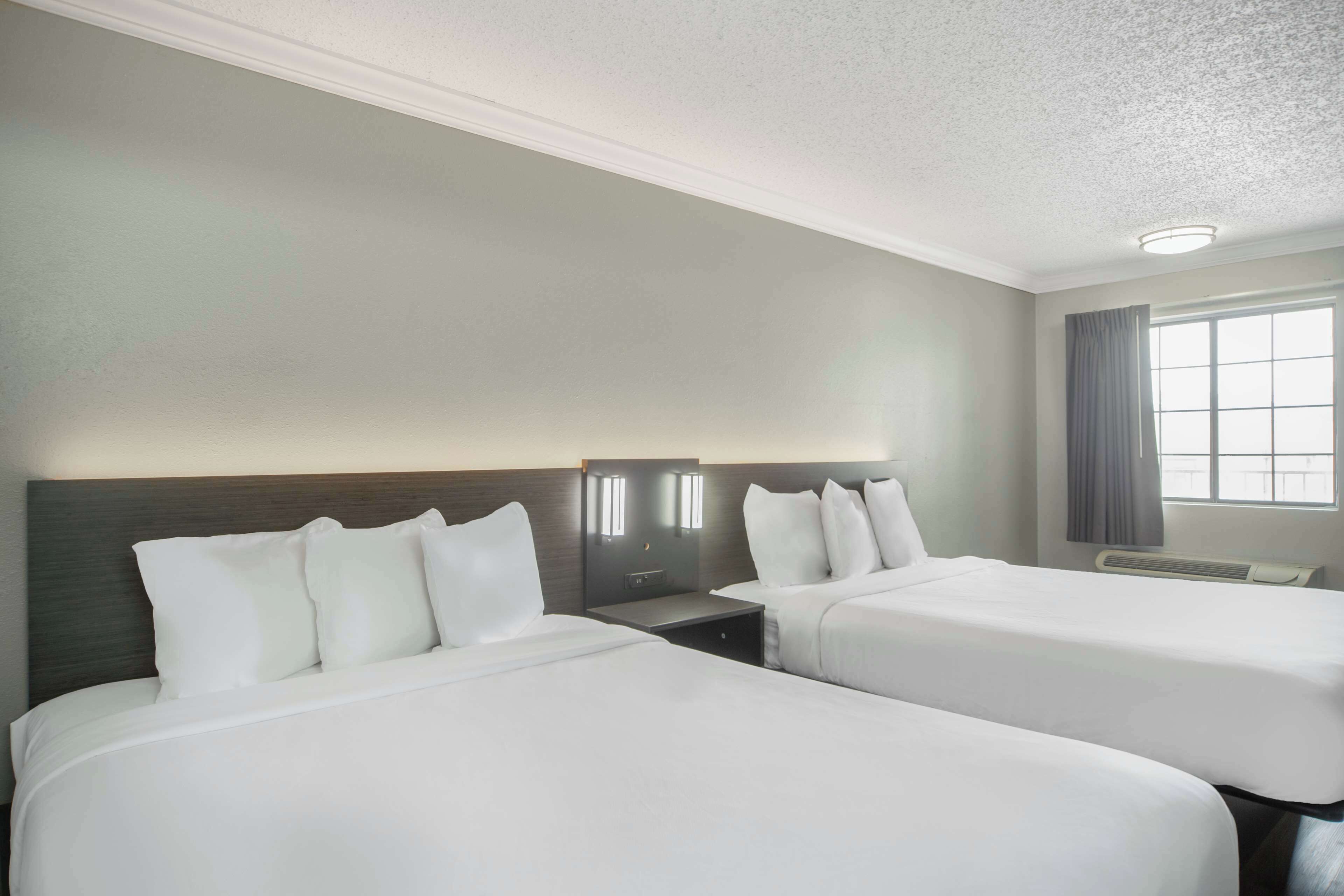Habitación de hotel con dos camas, una mesita de noche, una lámpara de pared y una ventana con cortinas.