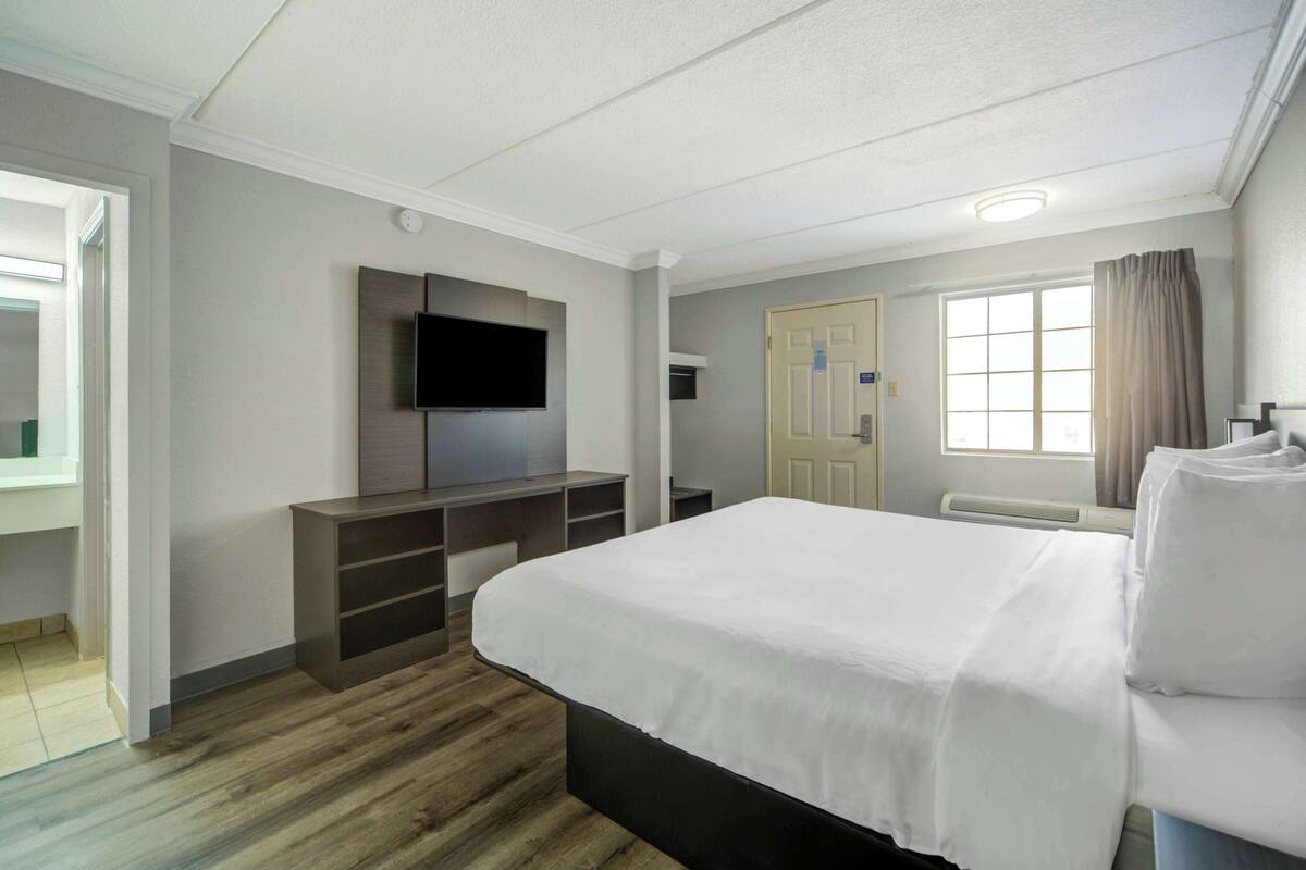 Ein Hotelzimmer mit einem großen Bett, einem flachbildfernseher an der Wand, einer eingebauten Ablage und einem fenster mit Vorhängen.