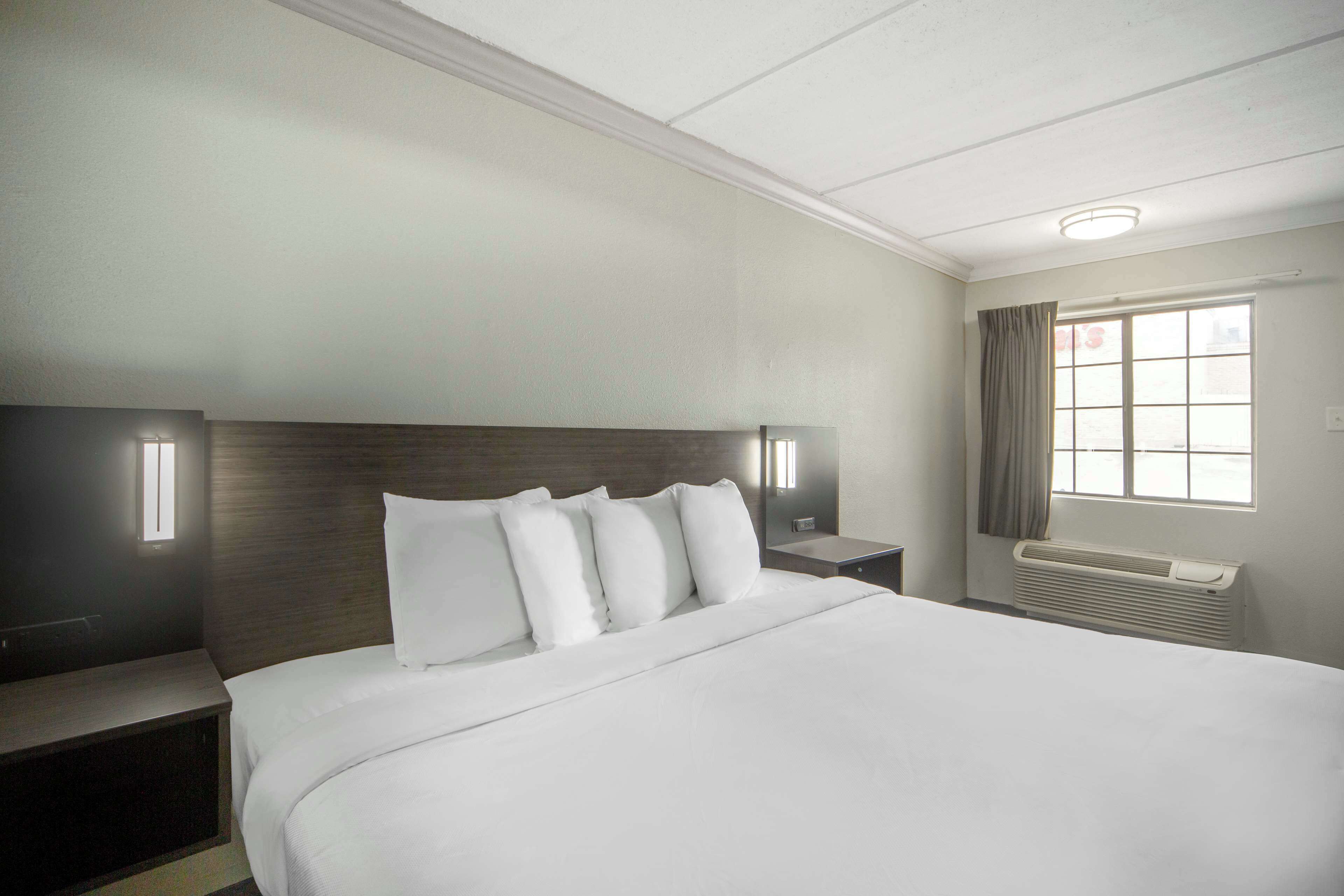 A hotel room with a large bed, a nightstand, a wall-mounted lamp, and a window with curtains.