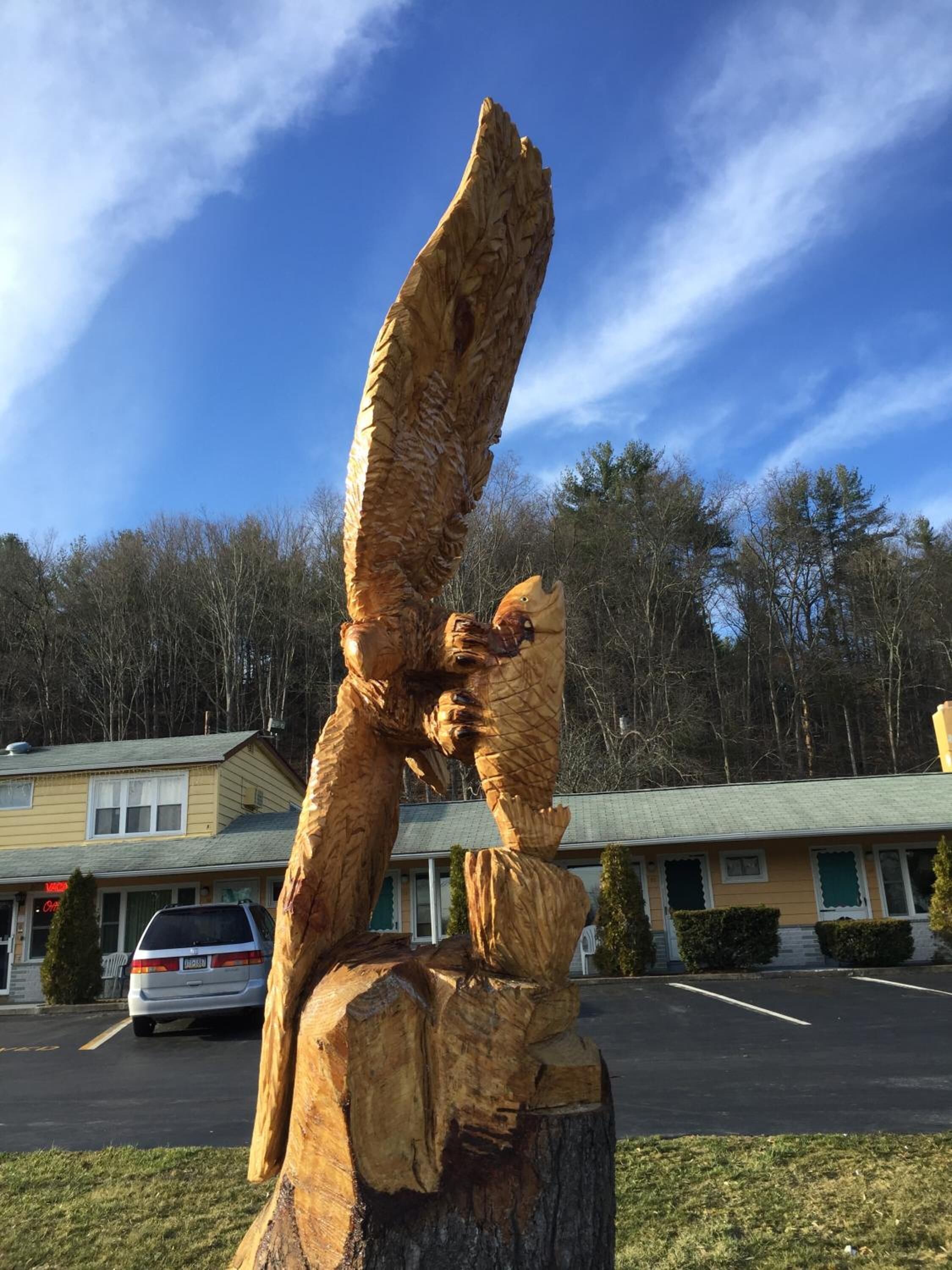 Una escultura de madera de un ángel en un estacionamiento, con casas al fondo.