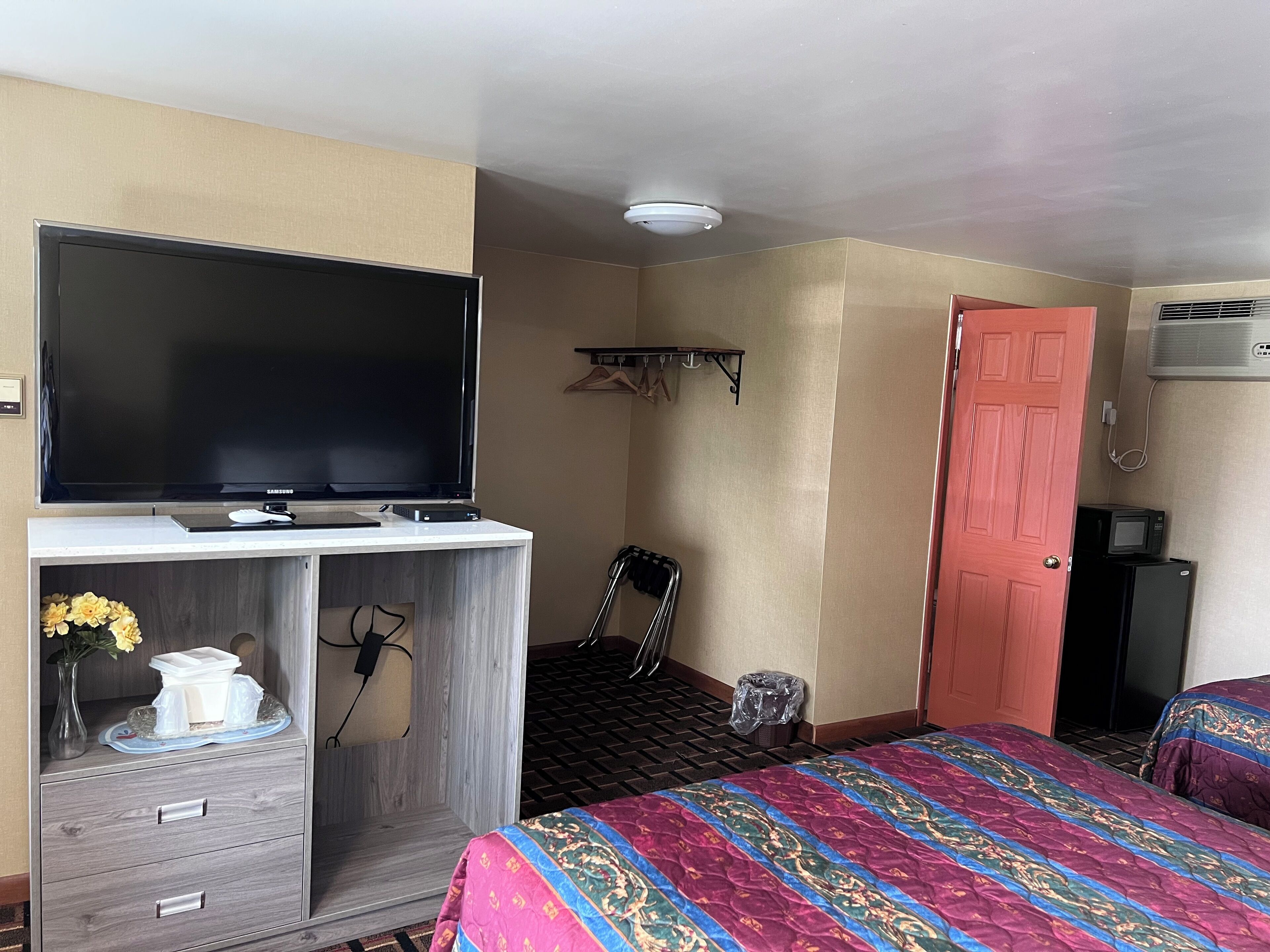 A hotel room with a flat-screen TV on a wooden cabinet, a bed with a colorful quilt, a chair, a small trash bin, and a red door.