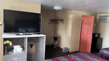 A hotel room with a flat-screen TV on a wooden cabinet, a bed with a colorful quilt, a chair, a small trash bin, and a red door.