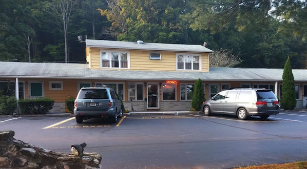 Um motel de dois andares com um letreiro de 'VACANCY', estacionamento e um cenário com árvores.