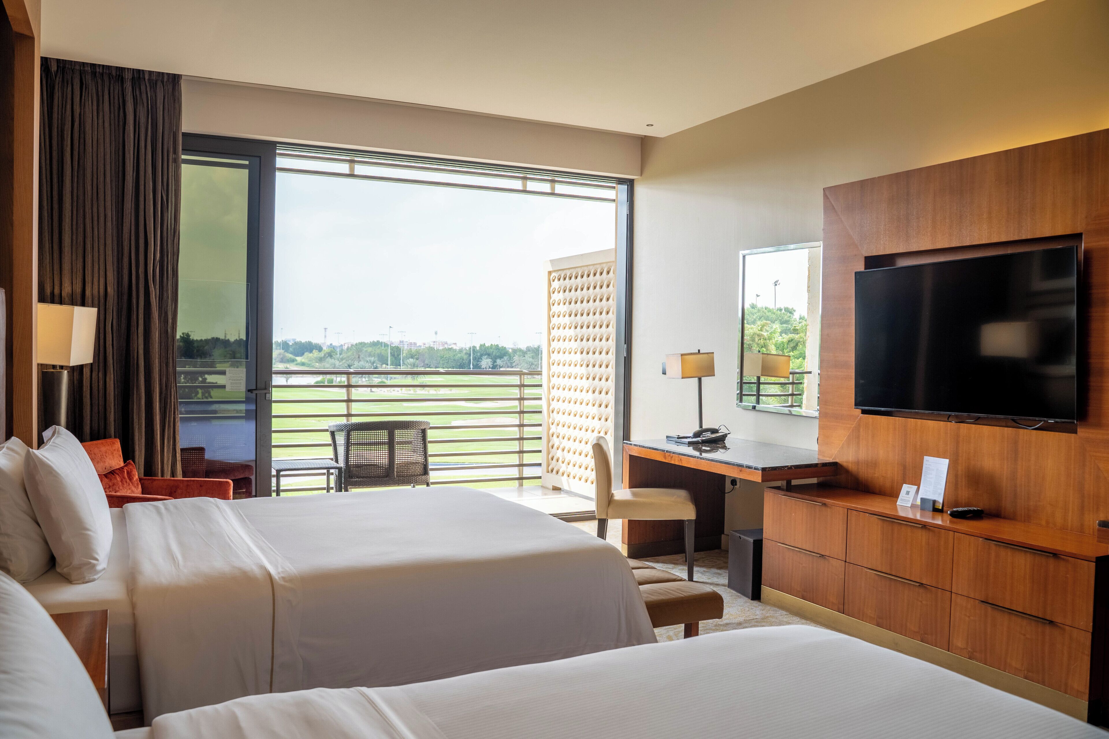 A hotel room with two beds, a desk, a TV, and a view of a green landscape.