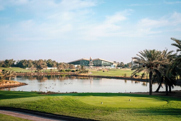 A golf course with a pond, palm trees, and a modern building in the background.