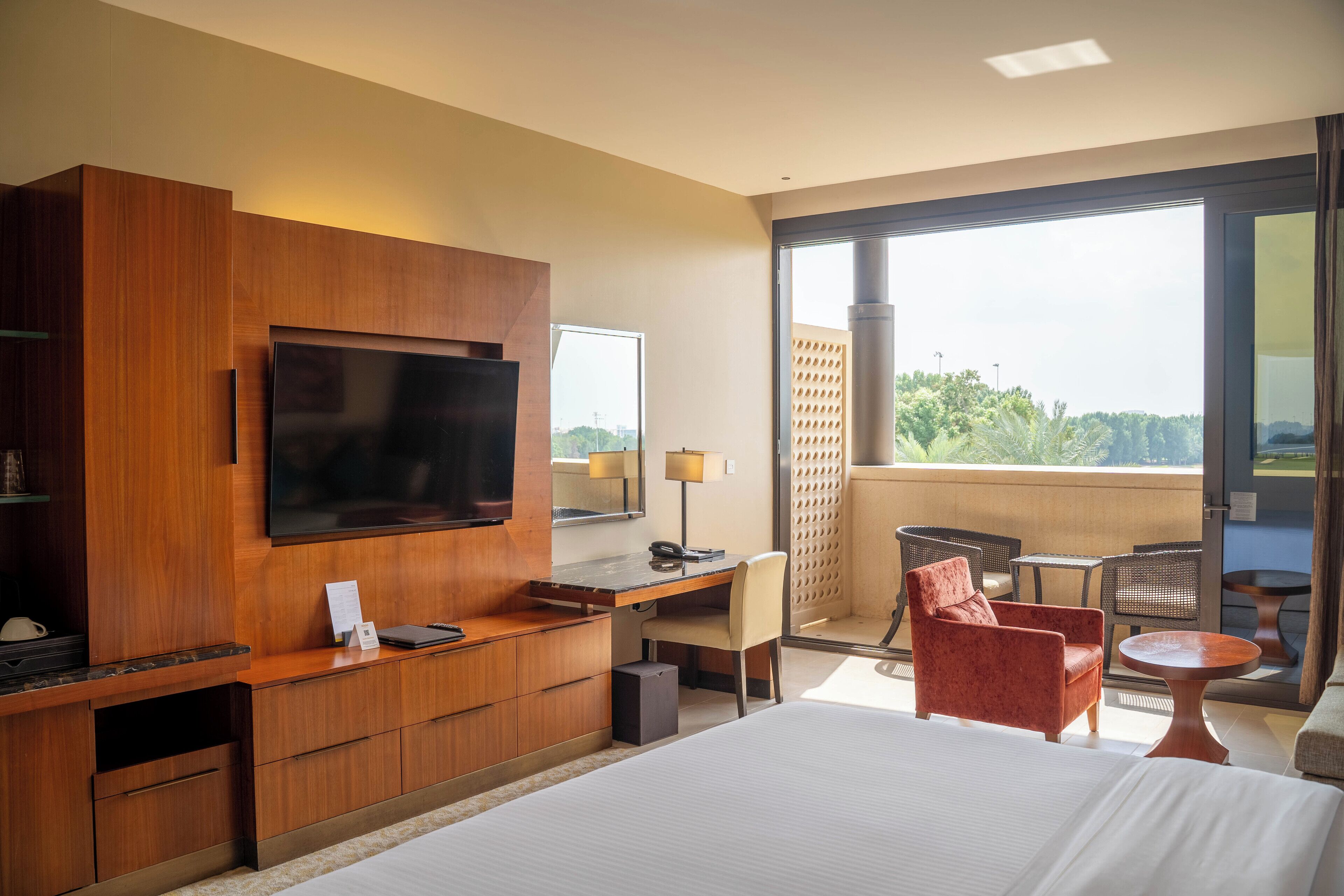 A hotel room with a large bed, a TV mounted on a wooden cabinet, a desk with a chair, a small table, and a balcony with chairs and a table.