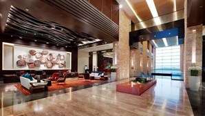 A spacious lobby with a modern design, featuring a large abstract art piece on the wall, a seating area with sofas and tables, and a large window providing natural light.