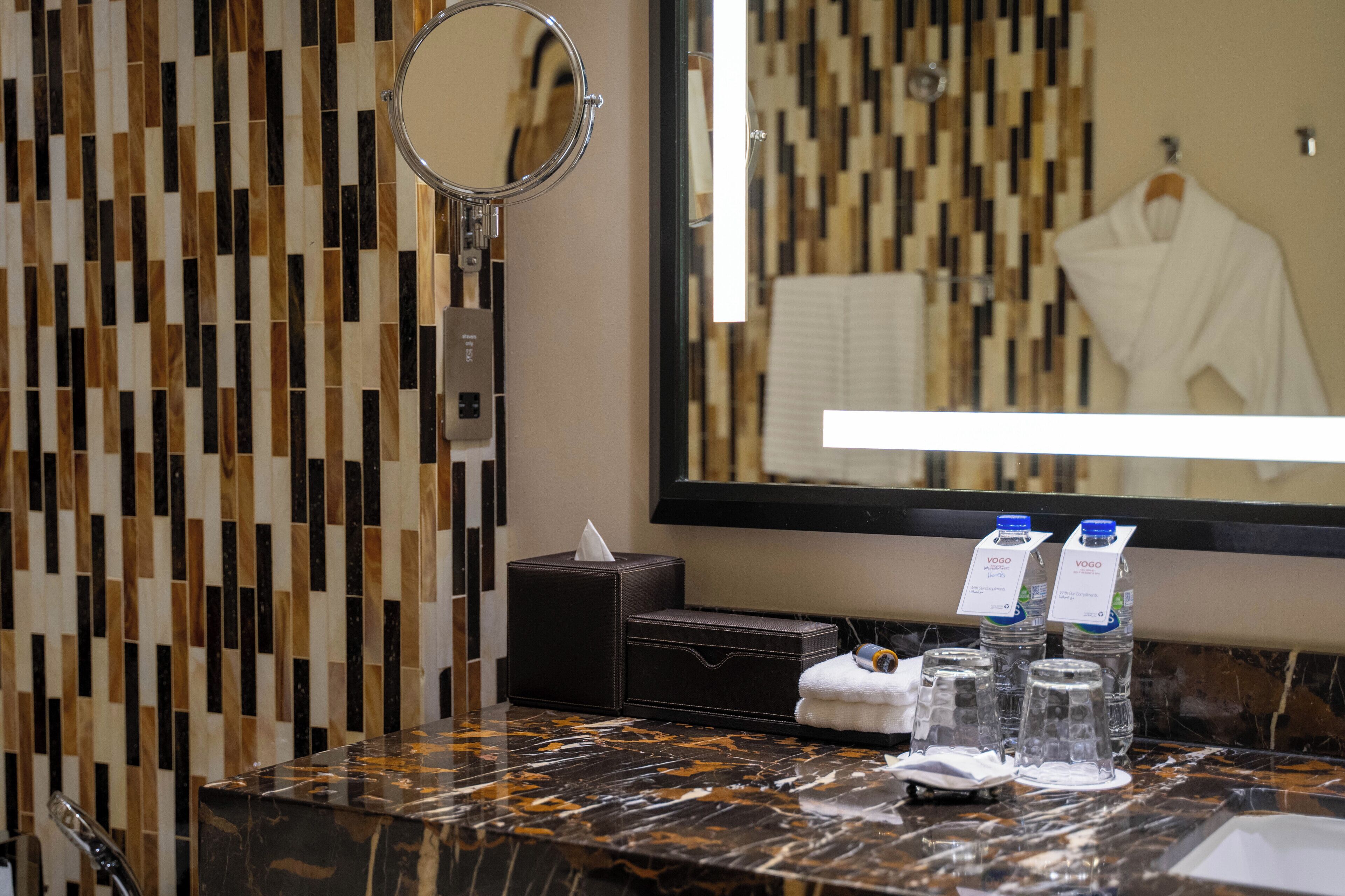 A bathroom with a marble countertop, a towel, a tissue box, a mirror, and VOGO water bottles.