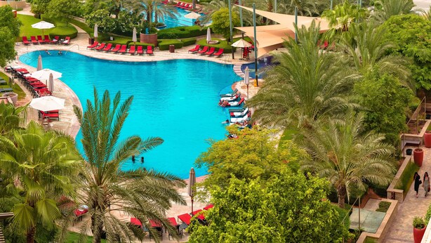 Ein großer Pool, umgeben von roten Liegestühlen und Sonnenschirmen, mit Palmen und einem kleinen Wasserfall.