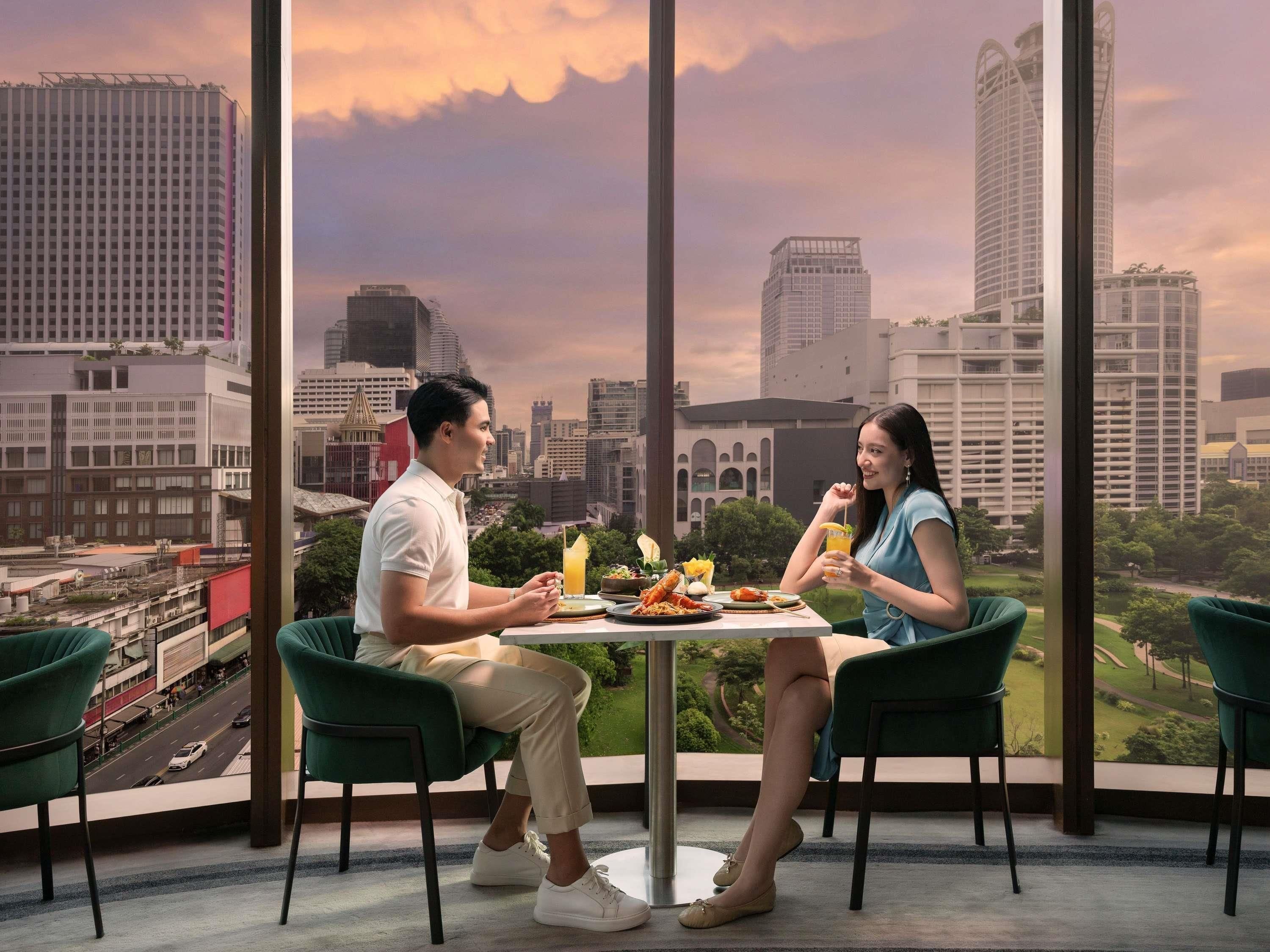 A dining area with a city view, two people seated at a table, and a table with food and drinks.
