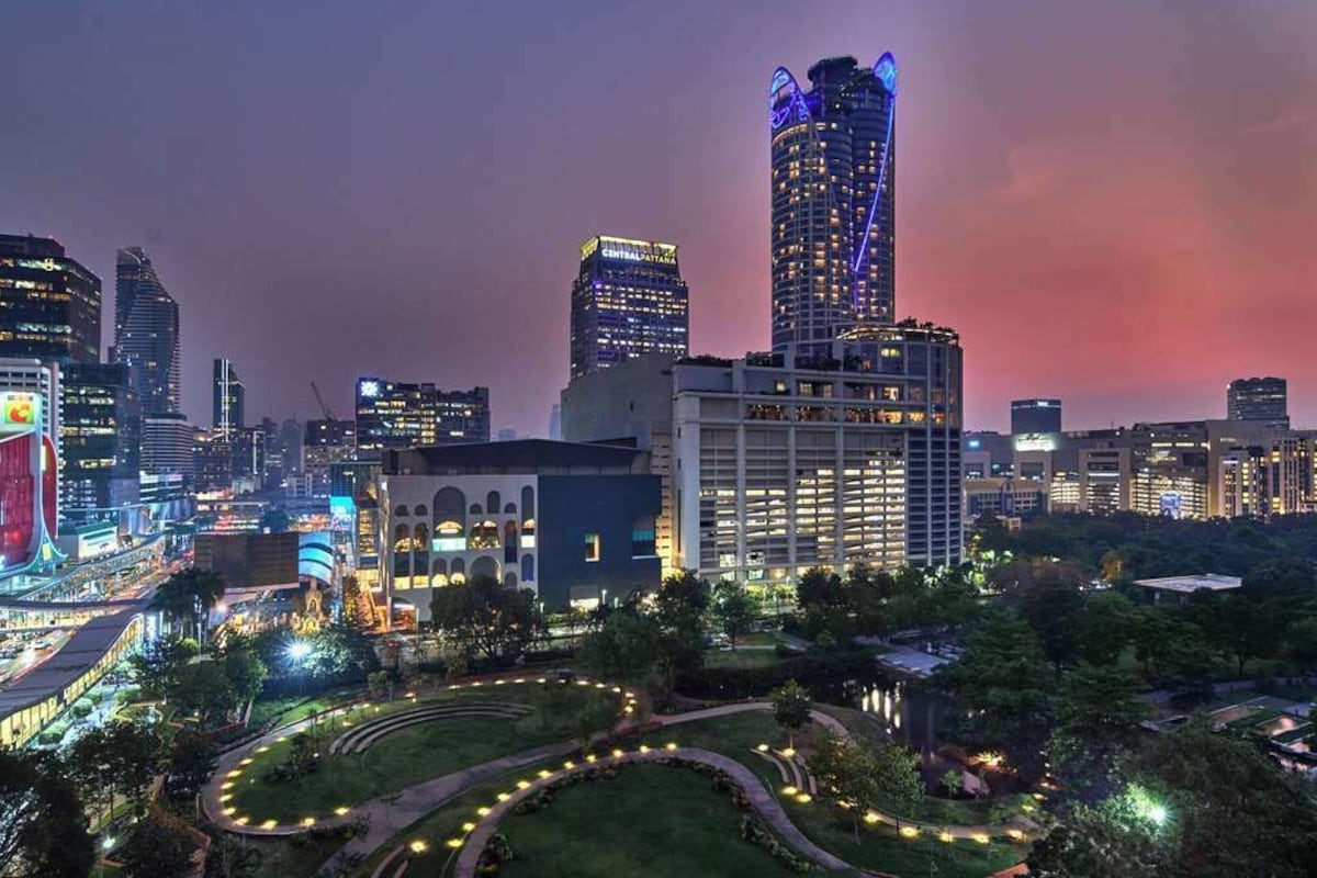 A cityscape at dusk with illuminated buildings, a prominent hotel with a lit sign, and a landscaped area with pathways.