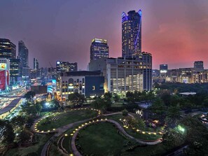 A cityscape at dusk with illuminated buildings, a prominent hotel with a lit sign, and a landscaped area with pathways. - Novotel Bangkok Platinum Pratunam (Bangkok)