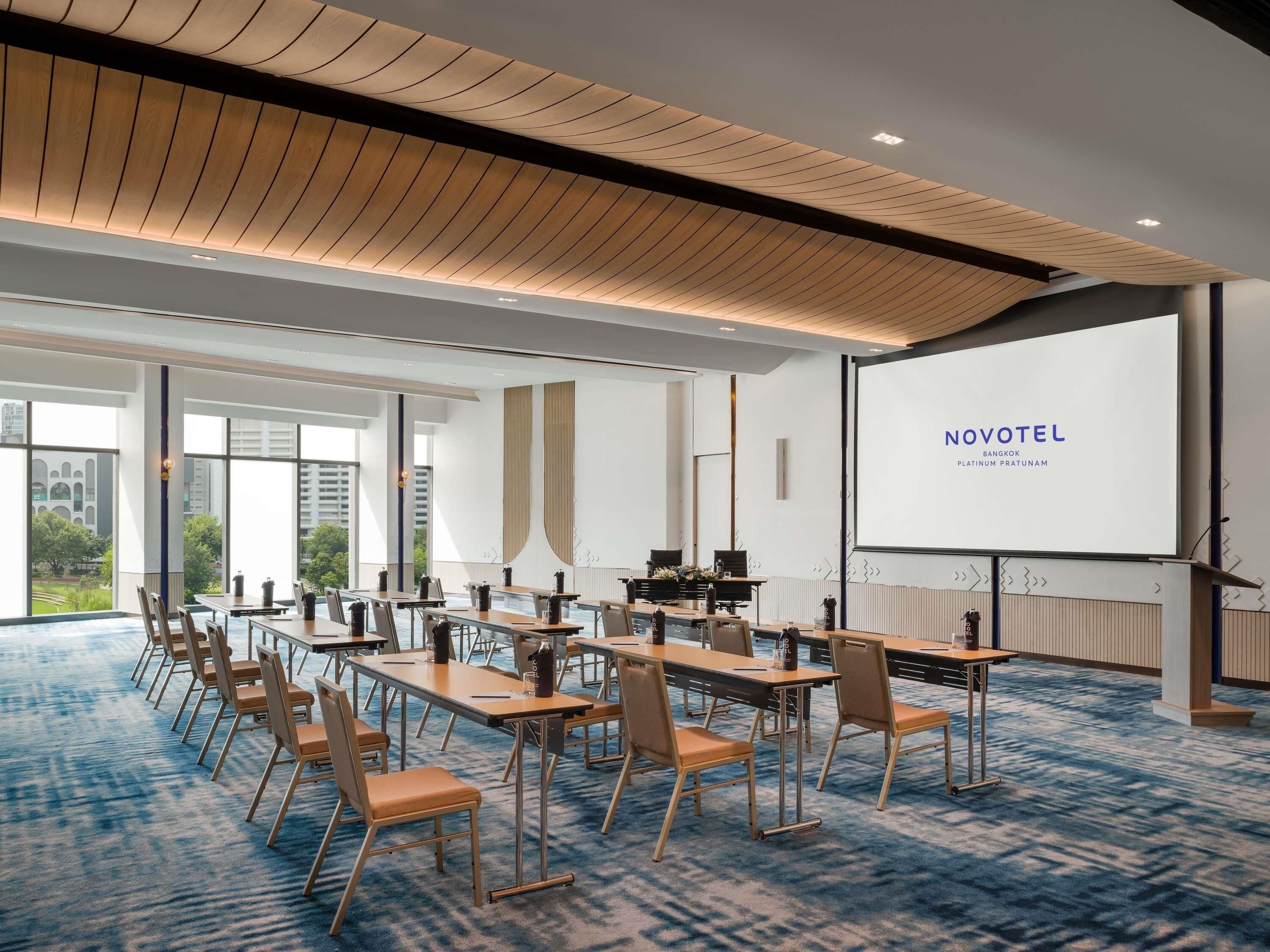 A modern hotel conference room with a large screen displaying 'NOVOTEL Bangkok Platinum Pratunam.'