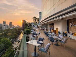 An outdoor dining area with tables and chairs, overlooking a cityscape and a river. - Novotel Bangkok Platinum Pratunam (Bangkok)