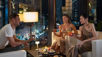 Quarto de hotel moderno com vista para a cidade, uma mesa com bebidas e petiscos, e uma luminária.
