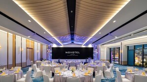Una sala conferenze con tavoli rotondi, sedie bianche e un grande schermo che mostra 'NOVOTEL BANGKOK PLATINUM PRATUMAN'.