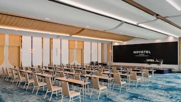 A conference room with rows of tables and chairs, a stage with a screen displaying 'NOVOTEL BANGKOK PLATINUM PRATUNAM', and a blue patterned carpet.