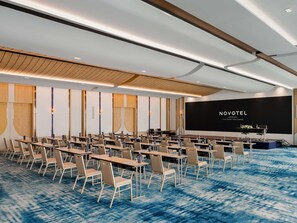 A conference room with rows of tables and chairs, a stage with a screen displaying 'NOVOTEL BANGKOK PLATINUM PRATUNAM', and a blue patterned carpet. - Novotel Bangkok Platinum Pratunam (Bangkok)